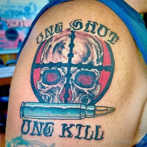 Фото тату на плече: череп в круге с надписью 'One Shot, One Kill' и патроном. Черно-синяя цветовая гамма, реалистичный стиль.