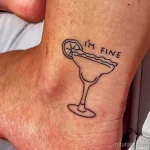 Фото тату на стопе с изображением бокала для мартини и надписью 'I'm Fine'. Простой, но выразительный рисунок.