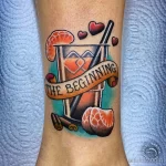 Фото тату красочного коктейля в бокале с апельсином, вишней и надписью "The Beginning". Стильный и оригинальный рисунок.
