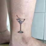 Фото тату минималистичного рисунка бокала мартини с оливкой на лодыжке. Черные контуры, простой дизайн, эскиз для любителей коктейлей.
