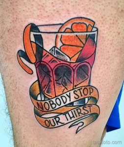 Яркое цветное фото тату с изображением коктейля в бокале и надписью "Nobody Stop Our Thirst". Татуировка выполнена в стиле олдскул.