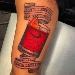 Фото тату на руке с изображением стакана Negroni, украшенного апельсиновой цедрой, и итальянской цитаты, выполненное в реалистичном стиле.