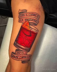 Фото тату на руке с изображением стакана Negroni, украшенного апельсиновой цедрой, и итальянской цитаты, выполненное в реалистичном стиле.