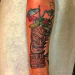 Фото тату на предплечье: красочная Тики-кружка с розовым фламинго, зонтиком и экзотическими фруктами. Идеальный эскиз тату для любителей тропиков.