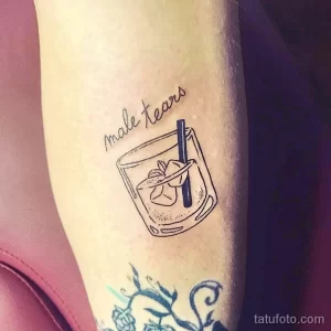 Фото тату на предплечье: стакан с коктейлем и надписью "male tears". Минималистичный стиль и тонкие линии.