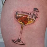 Фото тату: реалистичный рисунок бокала с красным напитком, внутри которого находится человек, погруженный в раздумья, и череп.