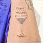 Фото тату: минималистичный рисунок бокала для мартини с надписью 'FUNKY COLD MEDINA' на предплечье. Черно-серая татуировка, выполненная тонкими линиями.