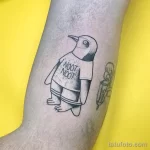 Фотография татуировки с изображением пингвина в футболке с надписью "NOOT NOOT" на ноге. Рисунок выполнен в черно-белых тонах.