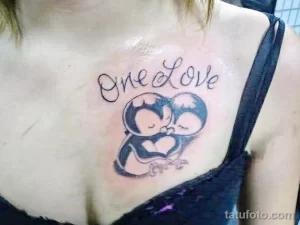 Фото татуировки двух пингвинов, обнимающих друг друга, с надписью "One Love" над ними, выполненной в черных чернилах.