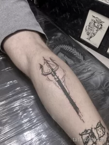 Фото тату: простой, но эффектный рисунок трезубца Посейдона в технике blackwork на икре, лаконичный дизайн.