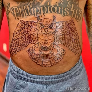 Фото большой татуировки совы и черепа с надписью "Philippians 4:13" на животе. Детализированный рисунок в черно-белых тонах.