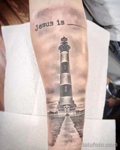 Фото реалистичной татуировки маяка на руке с надписью 'Jesus is...'. Детализированный рисунок в черно-серых тонах, выполненный с высокой точностью.
