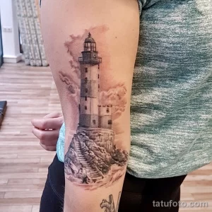 Фото тату: реалистичный рисунок маяка на предплечье, выполненный в черно-серых тонах с детализированными облаками и волнами.