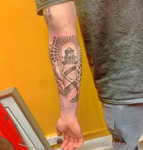 Фото татуировки на руке в технике black and grey с изображением маяка, окруженного лучами солнца и морскими волнами.
