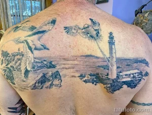 Фото тату на спине с изображением морского пейзажа, маяка, скал и летящих чаек. Выполнено в черно-серых тонах с детализированной проработкой.
