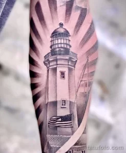 Фото татуировки маяка с развевающимся американским флагом на фоне лучей света, выполненной в реалистичном стиле.