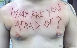 Фото тату: надпись "WHAT ARE YOU AFRAID OF?" выполнена техникой скарификации на груди мужчины. Видны следы свежих ран.
