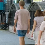 Фото тату на руке мужчины, идущего по улице. Виден небольшой рисунок на предплечье. Городской пейзаж и повседневная жизнь.