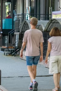 Фото тату на руке мужчины, идущего по улице. Виден небольшой рисунок на предплечье. Городской пейзаж и повседневная жизнь.