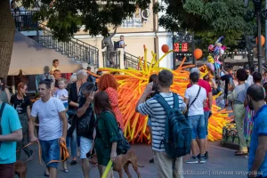 Фото тату: уличный артист в костюме из оранжевых и желтых воздушных шаров, окруженный прохожими на городской улице.