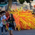 Фото тату: девушка проходит мимо уличного артиста в костюме из оранжевых и желтых воздушных шаров, окруженного зрителями.