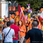 Фотография уличного шествия с людьми, держащими оранжевые шары, и огромной оранжевой медузой из воздушных шаров. Праздничная атмосфера и яркие краски.