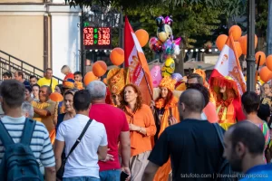 Фотография уличного шествия с людьми, держащими оранжевые шары, и огромной оранжевой медузой из воздушных шаров. Праздничная атмосфера и яркие краски.