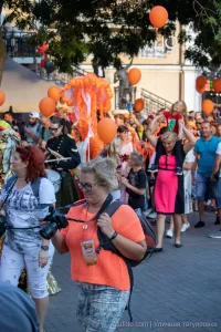 Фотография уличного шествия с фотожурналистом, снимающим оранжевую медузу из воздушных шаров. Динамичный момент и яркие краски.