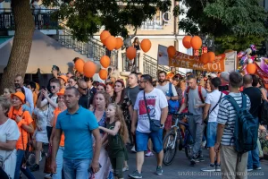 Фотография уличного шествия с множеством людей, держащих оранжевые шары. Праздничная атмосфера и яркие краски.