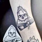 Фото тату на предплечье: изображение черепа, лежащего на открытой книге, с горящей свечой. Черно-белый рисунок в стиле реализм.