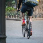 Фотография курьера Glovo на велосипеде с видимой татуировкой на ноге, едущего по улице. Курьер в шортах и носках, с желтым термо-рюкзаком.