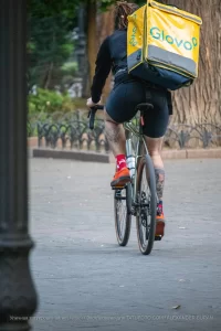 Фотография курьера Glovo на велосипеде с видимой татуировкой на ноге, едущего по улице. Курьер в шортах и носках, с желтым термо-рюкзаком.
