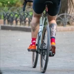 Фотография курьера Glovo на велосипеде с видимой татуировкой на ноге, едущего по улице. Курьер в шортах и носках, с желтым термо-рюкзаком.