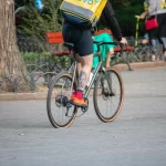 Фотография курьера Glovo на велосипеде с видимой татуировкой на ноге, едущего по улице. Курьер в шортах и носках, с желтым термо-рюкзаком.