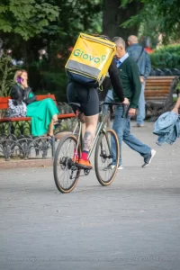 Фотография курьера Glovo на велосипеде с видимой татуировкой на ноге, едущего по улице. Курьер в шортах и носках, с желтым термо-рюкзаком.