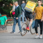 Фотография курьера Glovo на велосипеде с видимой татуировкой на ноге, едущего по улице. Курьер в шортах и носках, с желтым термо-рюкзаком.
