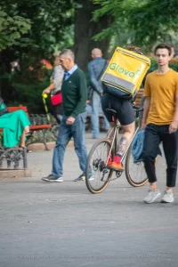 Фотография курьера Glovo на велосипеде с видимой татуировкой на ноге, едущего по улице. Курьер в шортах и носках, с желтым термо-рюкзаком.