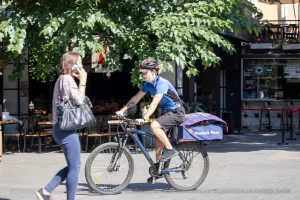 Фотография курьера на велосипеде с татуировкой на руке, едущего по городской улице. Курьер в синей форме и маске, разговаривает по телефону.