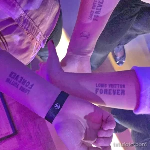 Фото парных татуировок 'Louis Vuitton Forever' на руках двух девушек в неоновом освещении. Татуировки выполнены черным цветом и выглядят стильно и современно.