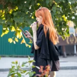 Фотография рыжеволосой девушки с татуировкой на руке, выпускающей пар от вейпа на фоне зелени и деревьев.