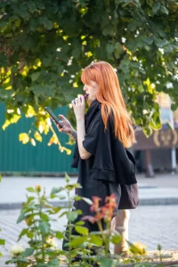 Фотография рыжеволосой девушки с татуировкой на руке, выпускающей пар от вейпа на фоне зелени и деревьев.