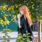Фотография рыжеволосой девушки с татуировкой на руке, выпускающей пар от вейпа на фоне зелени.