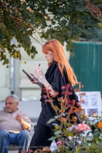Фотография рыжеволосой девушки с татуировкой на руке, держащей телефон и стоящей рядом с цветущими кустами.