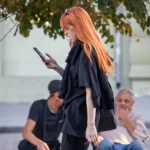 Фотография рыжеволосой девушки в черной одежде с татуировкой на руке, идущей по улице и выпускающей пар от вейпа.