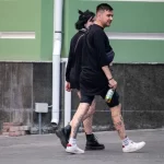 Фото тату на ноге девушки, идущей по улице. Рисунок выполнен в стиле стрит-арт, с использованием черных линий и небольших цветовых акцентов. Девушка одета в черную одежду и белые кроссовки.