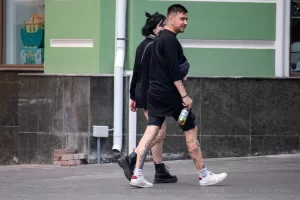 Фото тату на ноге девушки, идущей по улице. Рисунок выполнен в стиле стрит-арт, с использованием черных линий и небольших цветовых акцентов. Девушка одета в черную одежду и белые кроссовки.
