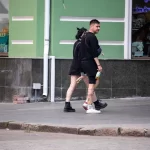 Фото тату на ноге мужчины, идущего по улице. Рисунок выполнен в стиле стрит-арт, с использованием черных линий и небольших цветовых акцентов. Мужчина одет в черную одежду.