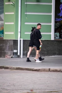 Фото тату на ноге мужчины, идущего по улице. Рисунок выполнен в стиле стрит-арт, с использованием черных линий и небольших цветовых акцентов. Мужчина одет в черную одежду.