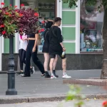 Фото тату на ноге девушки, идущей по улице. Рисунок выполнен в стиле стрит-арт, с использованием черных линий и небольших цветовых акцентов. Девушка одета в черную одежду.