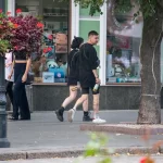 Фото тату на ноге девушки, идущей по улице. Рисунок выполнен в стиле стрит-арт, с использованием черных линий и небольших цветовых акцентов. Девушка одета в шорты и сетчатые колготки.